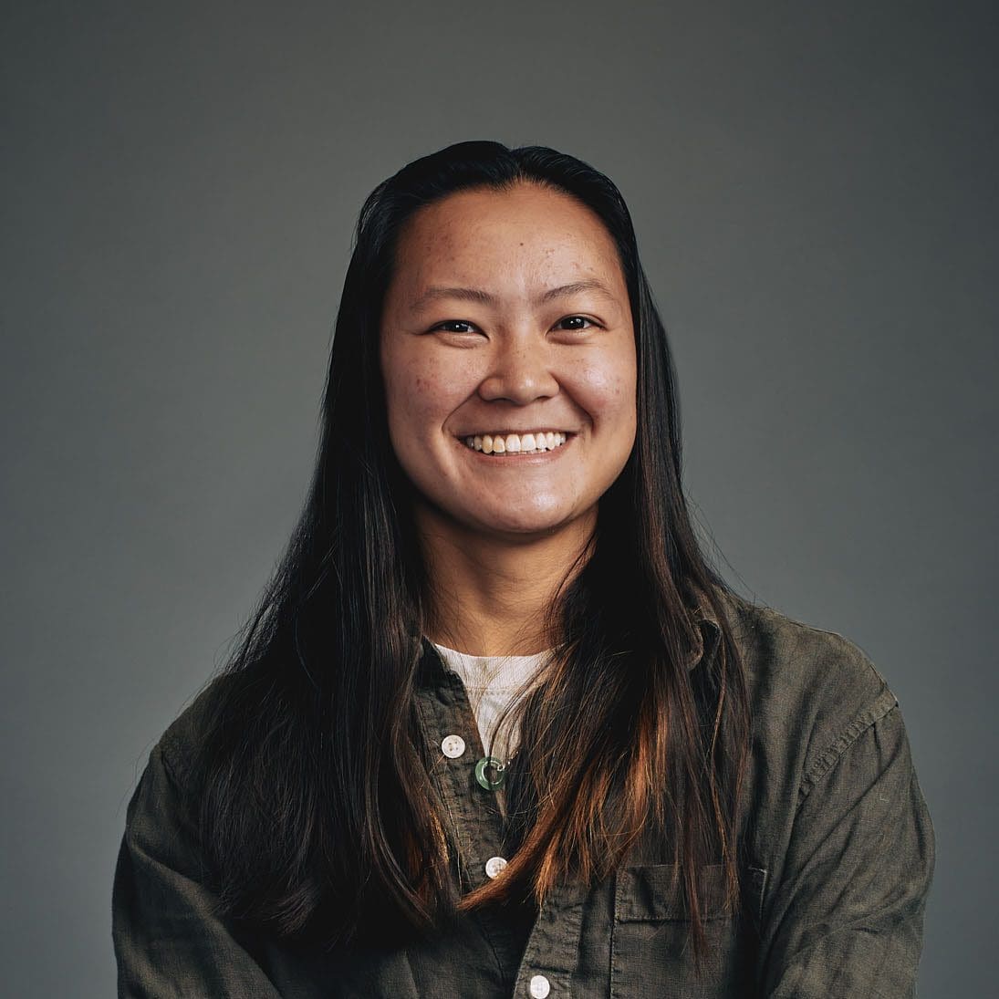 A young woman with long black hair smiles directly at the camera. She is wearing a dark green shirt.