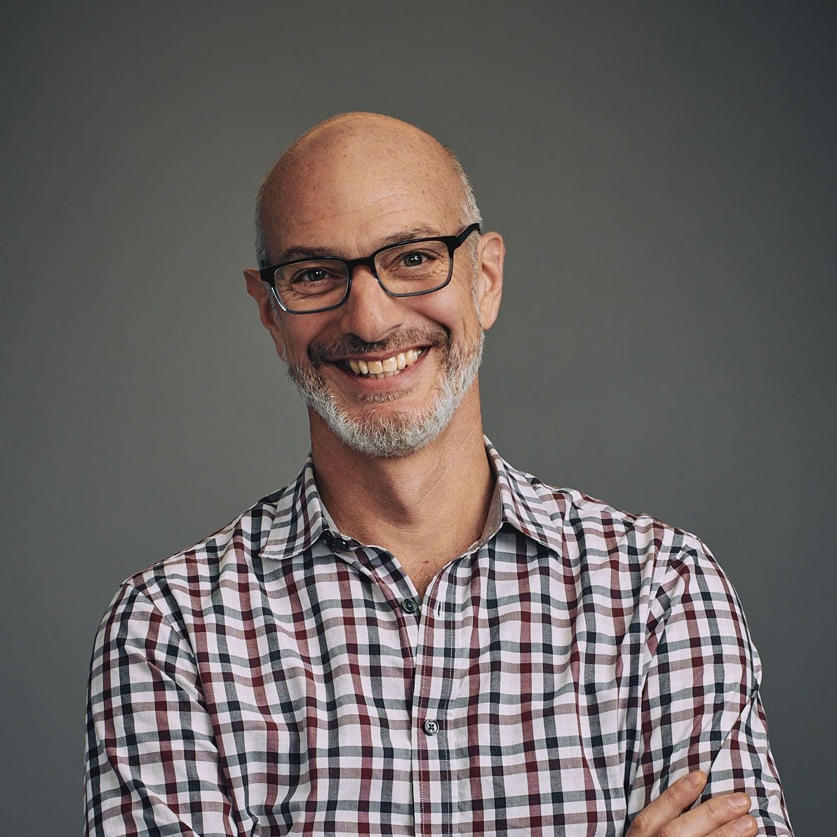 A tall man wearing glasses and a plaid shirt, smiles broadly at the camera. His head is shaved, and he has a beard. He is standing in front of a gray background.