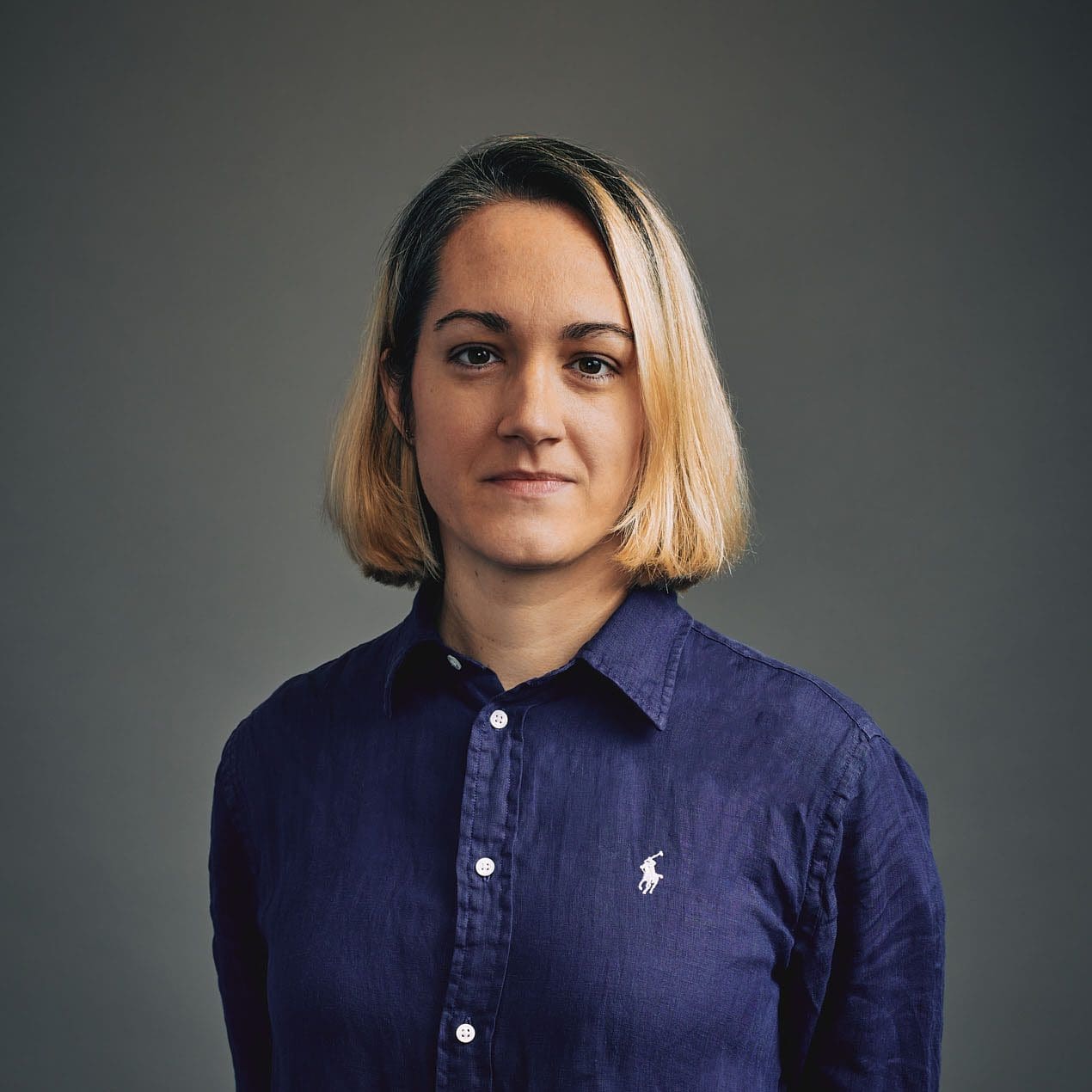 A white woman stares directly at the camera. Her hair is cut in a chin length bob and is blond with dark roots. She is wearing a blue button up shirt and is standing in front of a gray background.