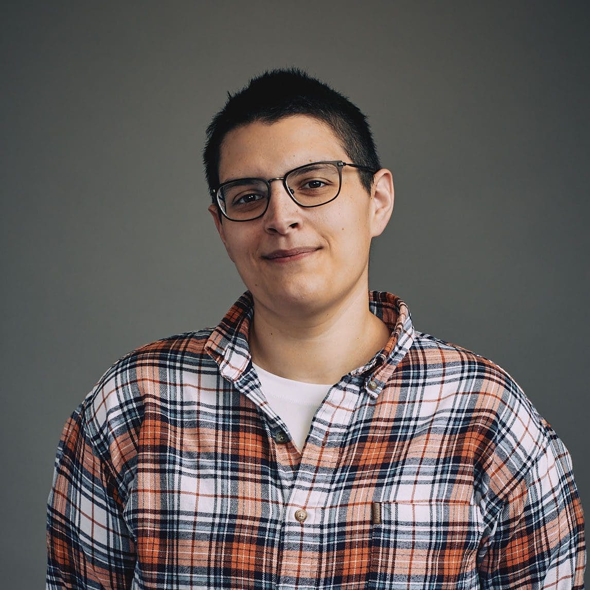 A non-binary individual with short black hair and glasses is half smiling at the camera. They are wearing a white and orange plaid shirt and standing with their hands behind them in front of a gray wall.