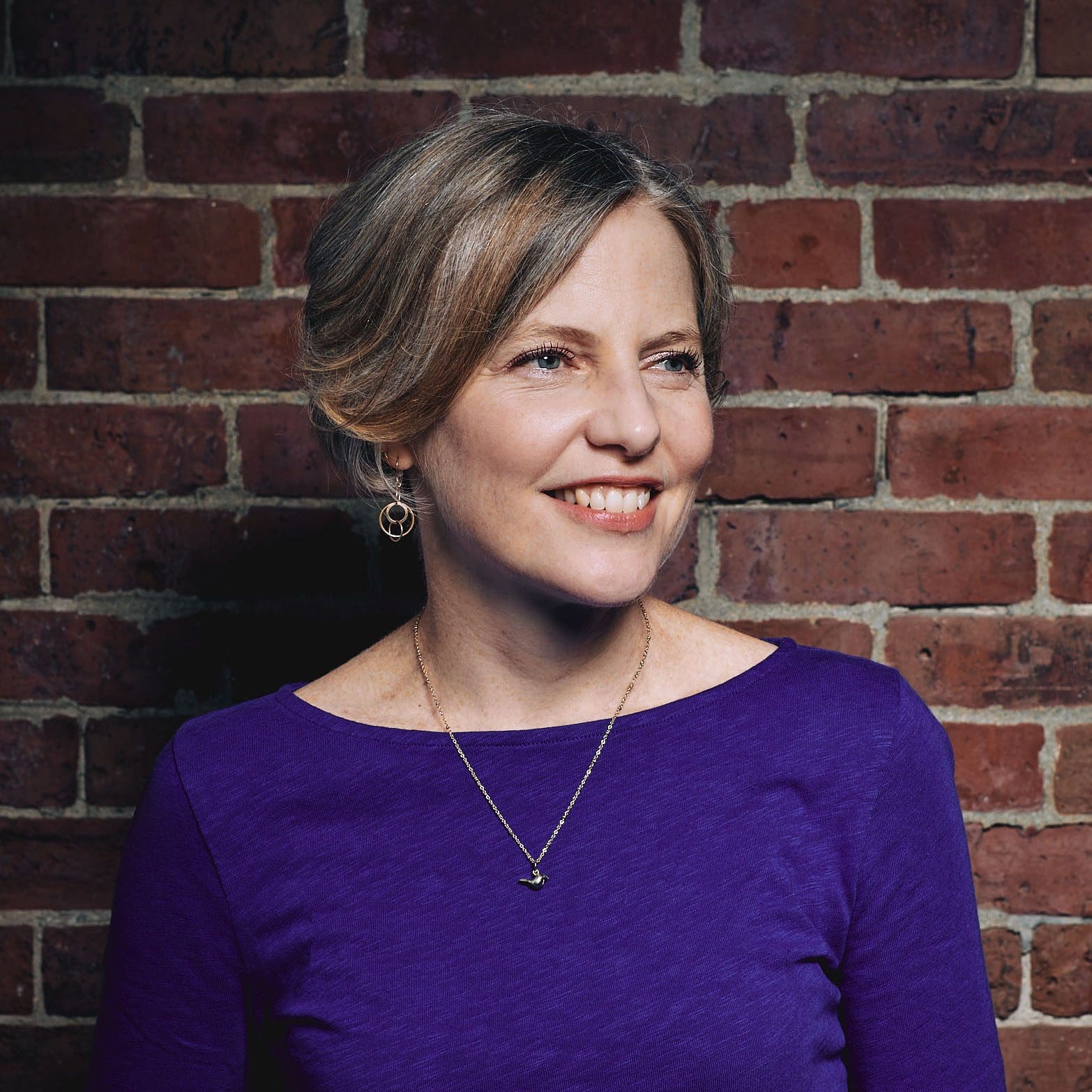 A woman with short dark blond hair smiles while looking away from the camera and to the right. She is wearing a royal blue shirt and standing in front of a brick wall.