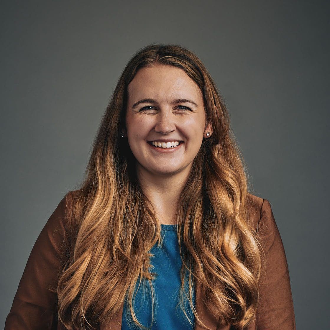 A young woman with long, wavy reddish blond hair. She has a big smile and is wearing a brown jacket over a turquoise-colored shirt in front of a gray background.