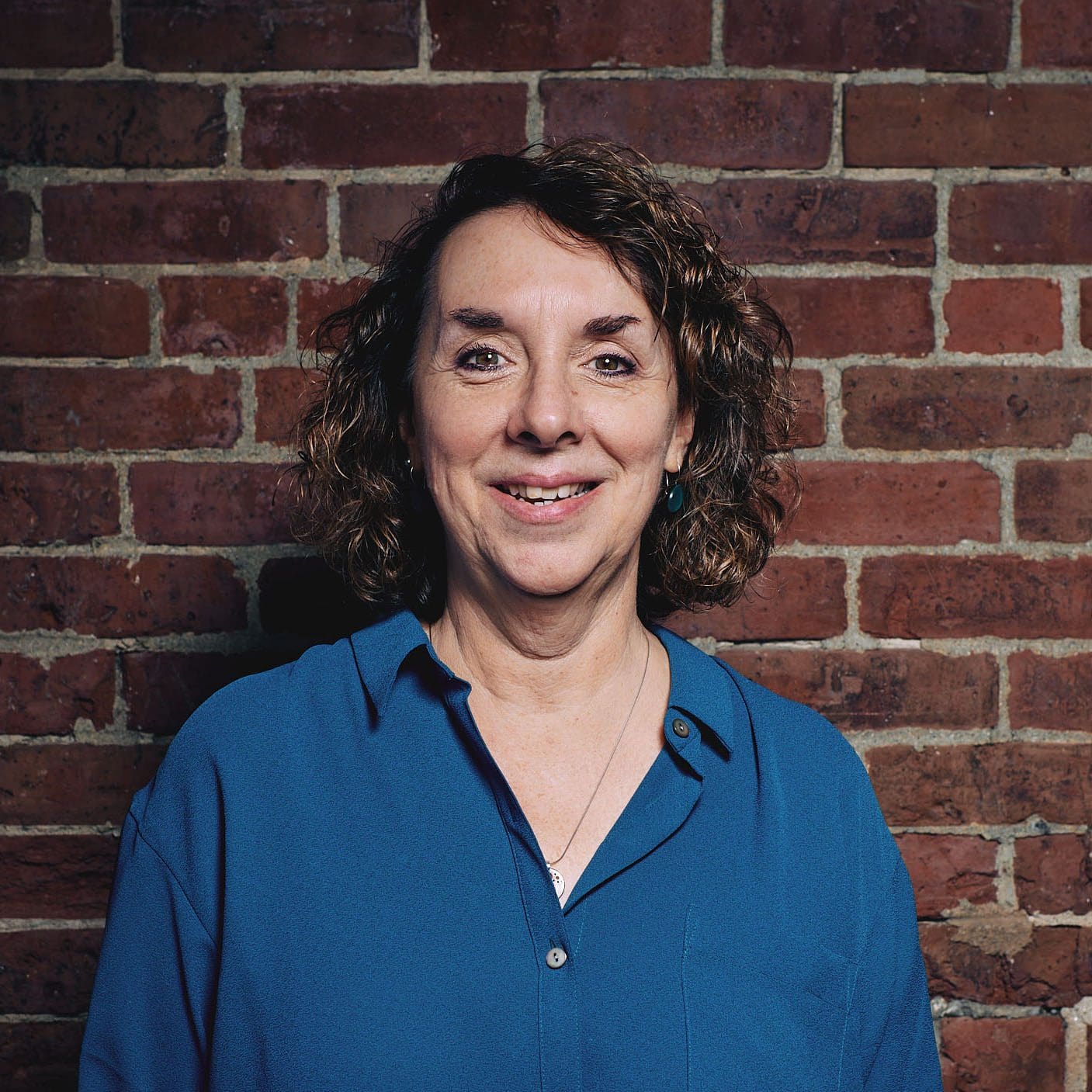 A mature woman with curly brown hair gazes directly at the camera with a smile. She is wearing a blue/green shirt and standing in front of a brick wall.