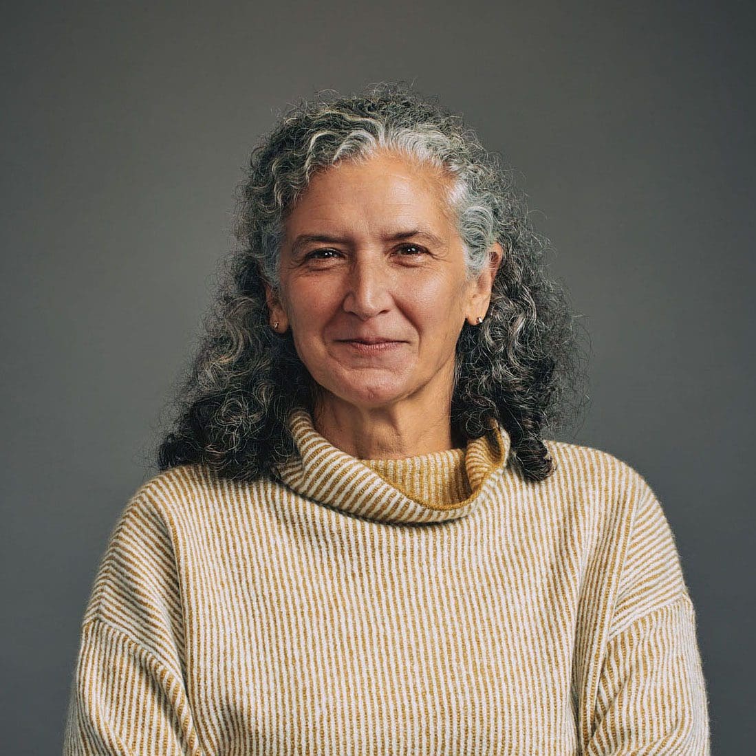 A woman with graying, curly hair stares directly at the camera with a half-smile. She is wearing a yellow turtleneck sweater and her arms are folded in front of her.