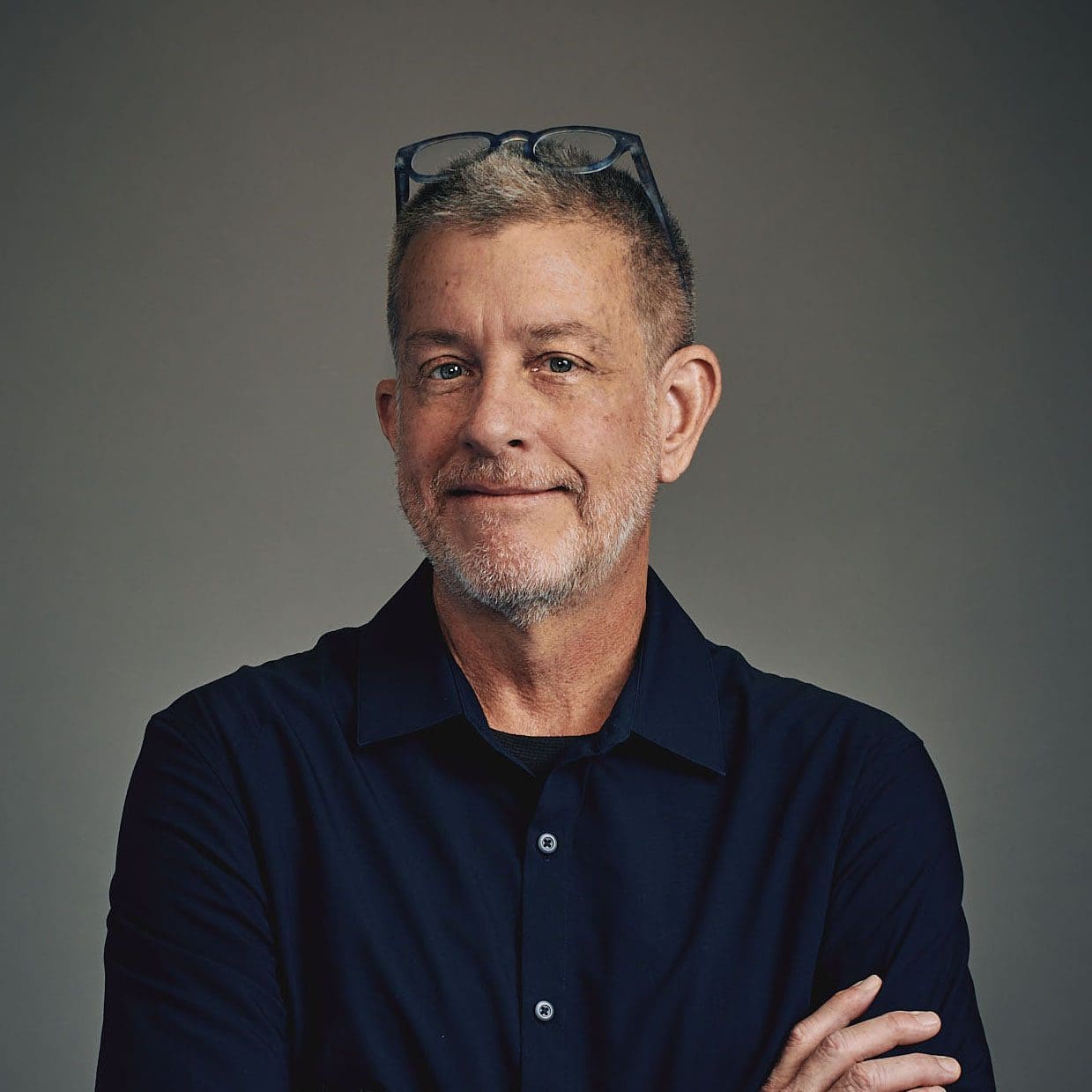 A man with graying blond hair wearing a navy-blue shirt with his glasses perched on his head stares directly at the camera. He is standing with his arms folded in front of a gray background.