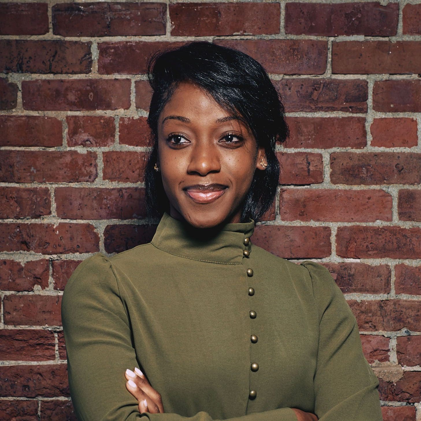 A woman with short, black hair gazes to the left with a slight smile. She is wearing an olive-green high collar shirt and her arms are folded in front of her. She is standing in front of a brick wall.