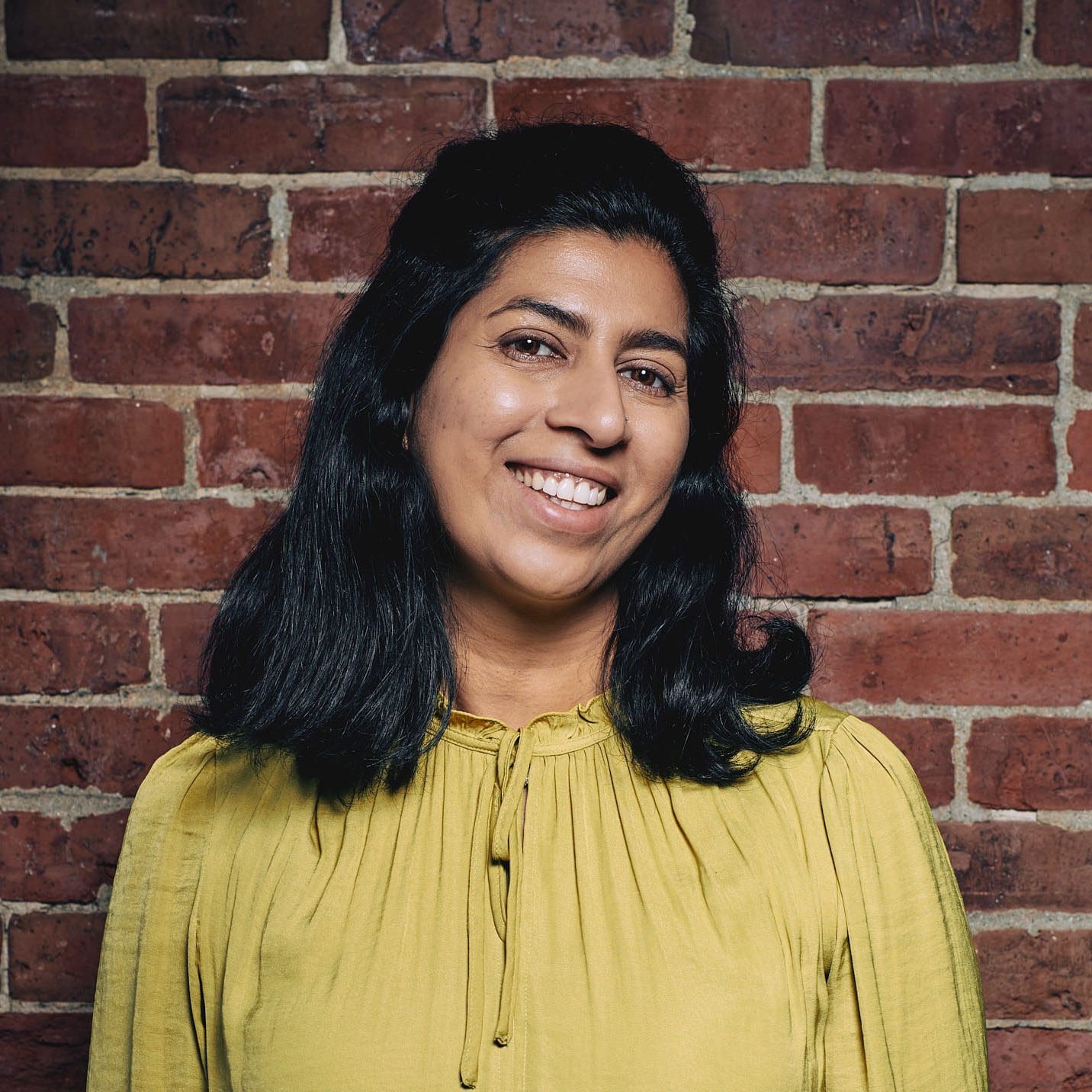 A woman with shoulder length black hair smiles at the camera. She is wearing a mustard yellow shirt and is standing in front of a brick wall.