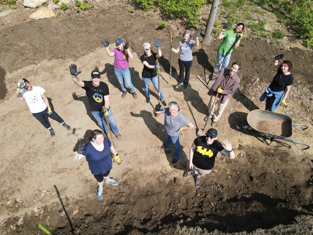 Ten KMA staffers in work gloves with shovels, rakes, and a wheel barrel in front of a large pile of dirt waving up at a drone camera.